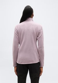 Άτομο με μακριά μαλλιά που φοράει ανοιχτό μωβ φλις μπουφάν με υφή και μαύρο παντελόνι, στέκεται γυρισμένο πλάτη σε απλό φόντο.