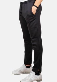Uomo che indossa pantaloni sportivi neri Adidas con cerniere laterali e sneaker bianche, in piedi con una mano in tasca contro uno sfondo neutro.