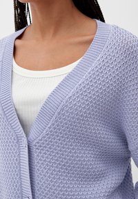 Světle fialový pletený cardigan s texturovaným vzorem, výstřihem do V a zapínáním na knoflíky. Nesený přes bílé žebrované tričko.
