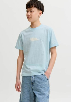 Tienerjongen met kort donker haar, gekleed in een lichtblauw t-shirt en losse lichte spijkerbroek, staat met één hand in de zak en kijkt naar links.