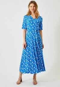 Blå blommig omlottdress med korta ärmar, ett knytmidja och lång kjol. Har ett livfullt mönster av vita blommor mot en blå bakgrund.