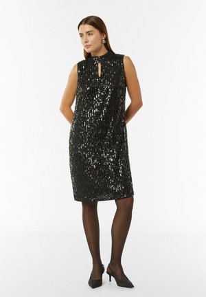 Robe sans manches noire avec un col haut, présentant un détail en forme de trou de serrure, recouverte de sequins. Associée à des collants foncés et des talons.
