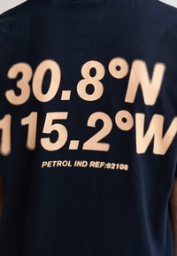 T-shirt en coton bleu marine avec un grand texte de couleur crème indiquant des coordonnées et "PETROL IND REF:92108" au dos.