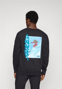 Man in een zwart sweatshirt met een kleurrijke afbeelding van een skiër in de lucht en grote blauwe "JAPED" tekst op de achterkant.