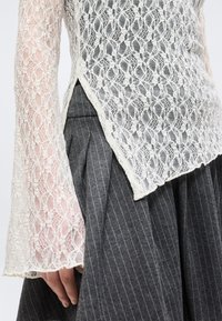 Λευκό τοπ μακρυμάνικο από δαντέλα με φλοράλ μοτίβο και κυματιστές άκρες, σε συνδυασμό με σκούρα γκρι πλισέ φούστα με ρίγες.