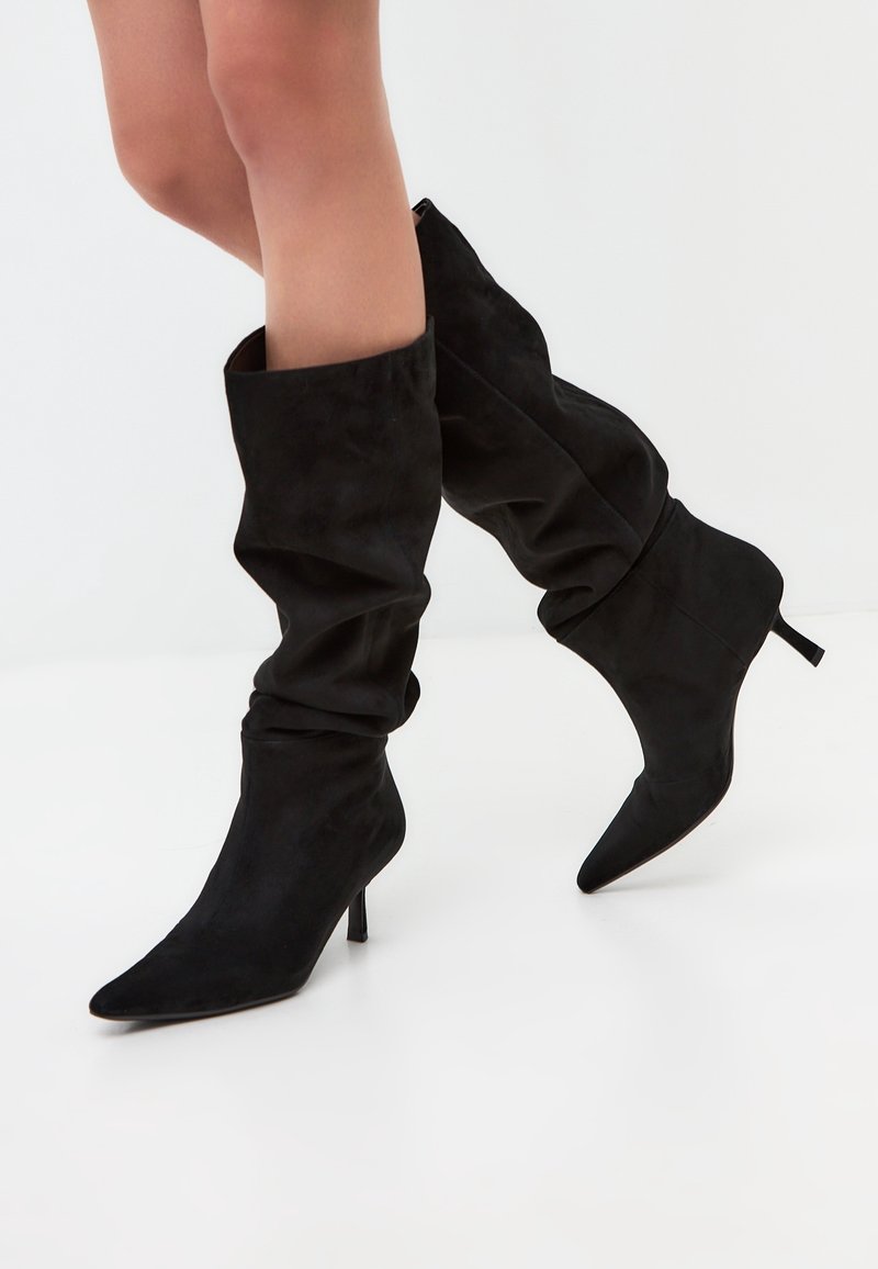 Botas negras de ante por encima de la rodilla con diseño holgado, punta afilada y tacón de aguja. La textura es suave y mate.