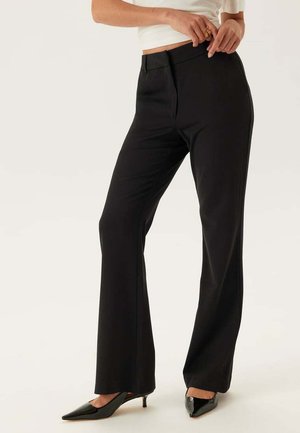 Vrouw in zwarte rechte broek, witte blouse die is ingestopt, en zwarte kitten heels, die de tailleband met haar handen aanpast.