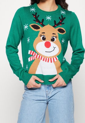 Groene trui met een glimlachend rendier dat een rood-wit gestreepte sjaal draagt, omringd door witte sneeuwvlokken, gedragen met lichtblauwe spijkerbroek.