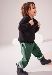 Hanorac negru cu glugă, pantaloni verzi largi cu bandă reflectorizantă la manșete și cizme negre din cauciuc pe un fundal deschis.