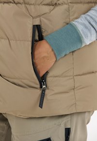 Beige Steppweste mit einer Reißverschlusstasche, die einen schwarzen Reißverschluss und einen Ziehgriff aufweist. Ein graues Langarmshirt schaut darunter hervor.