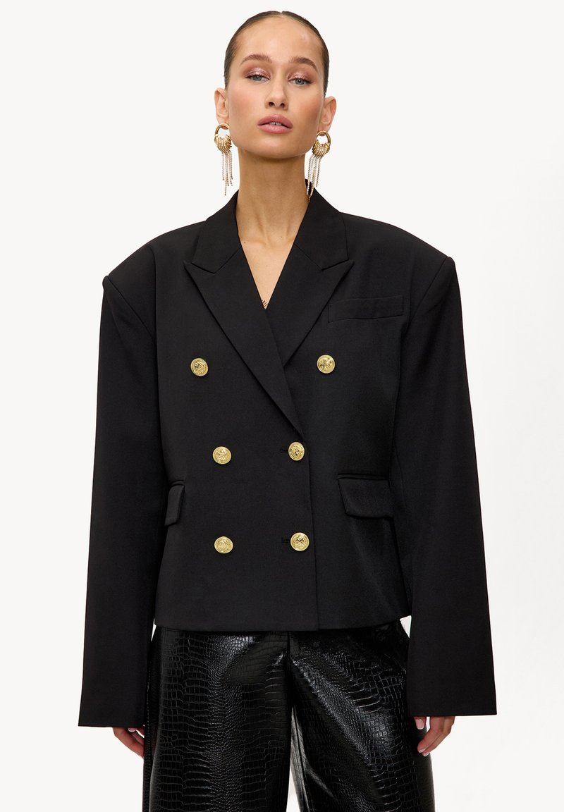 Blazer noir croisé à double boutonnage avec boutons dorés, épaules structurées et coupe courte ; associé à un pantalon en cuir verni noir brillant.