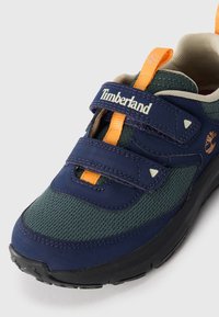 Marineblå og grønn Timberland sneakers med oransje draknapper og doble borrelåsremmer på svart såle, vinklet for å vise front- og sidestiler.