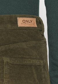 Olivově zelené manšestrové kalhoty s zadní kapsou a hnědou koženou etiketou našitou na pasu, na které je vytištěno "ONLY Blue Denim Since 1995".