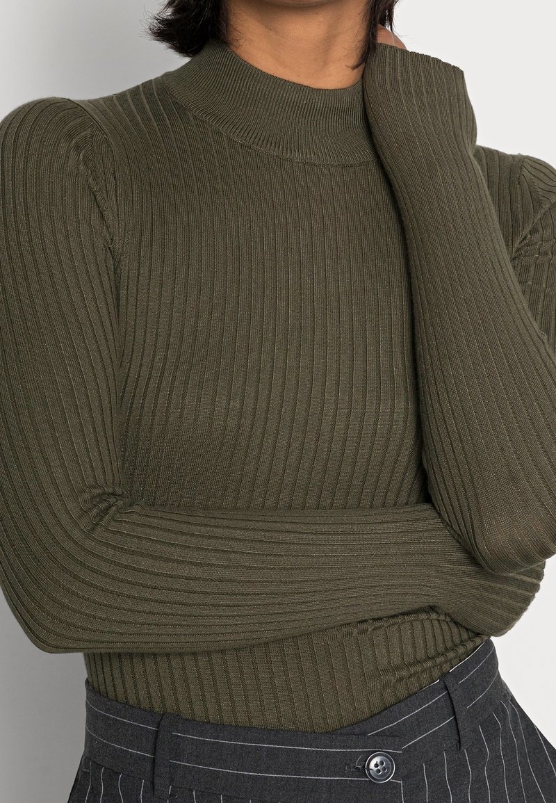 Personne portant un pull à manches longues côtelé vert olive et un pantalon gris à fines rayures, avec un bras croisé et la main touchant le cou.