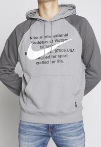 Grå och mörkgrå hoodie med en framficka, prydd med en vit Nike-logotyp och text. Tillverkad av mjukt tyg, med justerbar huva och ribbade ärmslut.