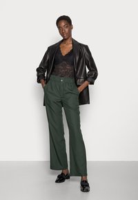 Top in pizzo nero, blazer in pelle nera oversize, pantaloni larghi verdi scuri con vita elasticizzata e chiusura frontale a bottone, mocassini neri.