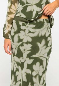 Mujer vestida con una falda y una blusa de patrón floral en verde y beige, con una mano levantando ligeramente la blusa.