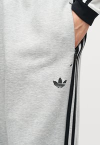 Main dans la poche d'un pantalon de survêtement Adidas gris clair avec des détails trois bandes noirs et un logo trèfle noir près de la cuisse.