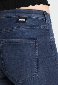 Vaqueros de mezclilla azul oscuro con una vista de cerca del bolsillo trasero, que presenta una etiqueta de cuero y costuras cosidas en una tela suave y texturizada.