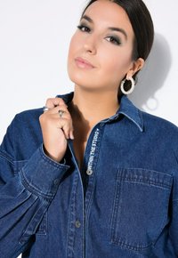 Camicia di jeans con colletto, maniche lunghe e taschino frontale. Colore blu scuro, leggera usura e dettagli di cucitura visibili.