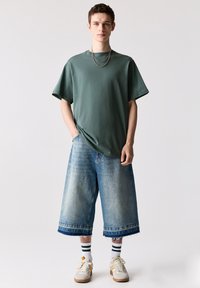 Overmaatse groene t-shirt gecombineerd met lichtblauwe denim culottes. Opvallende details zijn de rafelige zomen en witte sneakers met gele accenten.