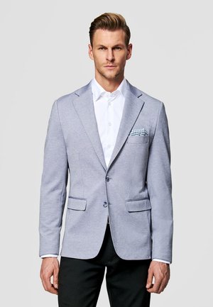 Mężczyzna ubrany w jasnoszary blezer na białej koszuli z ozdobną poszetką, stojący na jednolitym jasnym tle.