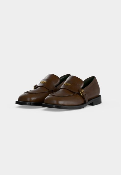 Paire de mocassins en cuir marron avec détails de boucles dorées et semelles noires, positionnés côte à côte sur un fond clair uni.