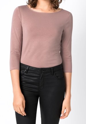 Femme portant un haut moulant mauve à manches longues trois-quarts et un pantalon noir taille haute sur un fond uni.