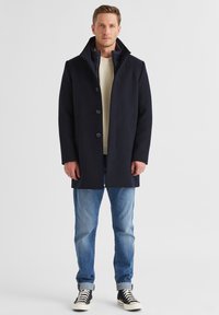 Marineblauer Wollmischungs-Mantel mit hohem Kragen, Fünf-Knopf-Verschluss, Seitentaschen und einer gesteppten Innenschicht, kombiniert mit blauen Jeans und Turnschuhen.