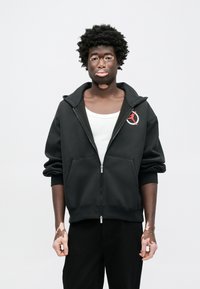 Μαύρη φούτερ με φερμουάρ και μεγάλο γιακά, δύο μπροστινές τσέπες και λογότυπο στο στήθος. Φορεμένη πάνω από λευκό αμάνικο τοπ και συνδυασμένη με μαύρο παντελόνι.