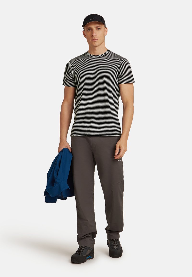 Pruhované černobílé tričko, šedé kalhoty a černé boty. Drží modrou bundu. Neformální design, krátké rukávy a jednoduchý vzor.