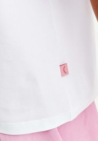 Bílý bavlněný tričko s zaobleným spodním okrajem a malou růžovou průsvitkou s jednoduchým designem řasy na boku. Měkká struktura, volný střih.