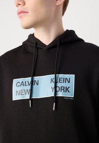 Zwarte hoodie met een grote blauwe grafische print met de tekst "CALVIN NEW KLEIN YORK." Voorzien van trekkoorden met zilveren uiteinden en een zachte textuur.
