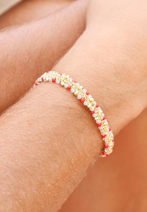 Bracelet ton or avec un design tissé comprenant des perles rouges et claires, affichant une apparence texturée sur un poignet bronzé.