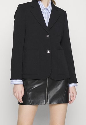 Kobieta ubrana w czarną dopasowaną marynarkę na niebieską koszulę w paski i czarną skórzaną mini spódnicę z przednim zamkiem.