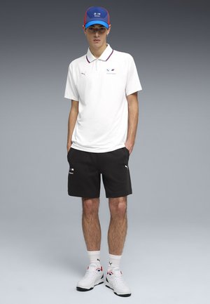Junger Mann trägt weißes BMW Motorsport Polo, schwarze Puma-Shorts, weiße Puma-Sneaker und blaue BMW Motorsport Kappe, steht mit Händen in den Taschen.