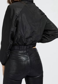 Chaqueta negra corta con una textura brillante y suave, con mangas largas y puños elásticos. Combinada con unos jeans ajustados y brillantes de color negro.