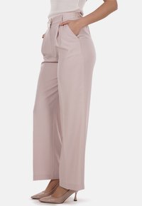 Pantaloni a gamba larga rosa chiaro con una texture liscia, vita alta, tasche frontali e un pannello posteriore elasticizzato. Indossati con tacchi beige.
