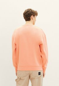 Luźna bluza z okrągłym dekoltem w jasnym koralowym kolorze, z długimi rękawami i ściągaczami, w zestawieniu z beżowymi spodniami cargo z widoczną metką.