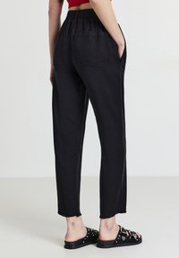 Personne portant un pantalon noir cropped avec une taille élastique et des ourlets effilochés, associé à des sandales à glissière noires cloutées et un haut rouge.