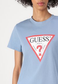 Hele helesinine puuvillane t-särk, millel on punane kolmnurkne logo kujundus, mille keskel on tekst "GUESS U.S.A. WASHED JEANS". Lühikeste varrukatega.