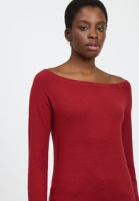 Maglione rosso in lana con scollo a spalla, maniche lunghe e texture liscia, caratterizzato da delicati dettagli delle cuciture sulle spalle.