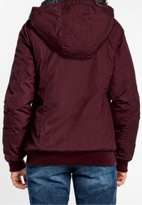 Kastanjefärgad vadderad jacka med huva, ribbat fåll och manschetter. Har en slät textur och en något glänsande yta. Blå denimjeans är synliga.