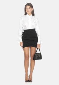 Camicia bianca a colletto, gonna nera aderente con design drappeggiato, borsa nera con logo geometrico, décolleté lucidi marroni. Tessuti morbidi.