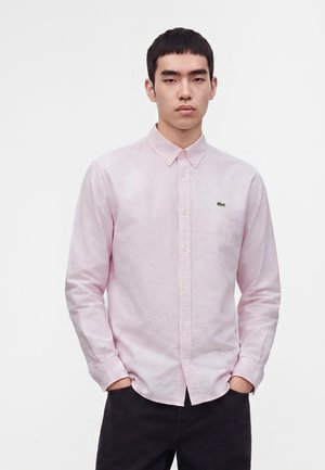 Giovane uomo che indossa una camicia rosa chiaro con bottoni e maniche lunghe e pantaloni neri, in piedi con le mani in tasca su uno sfondo semplice.
