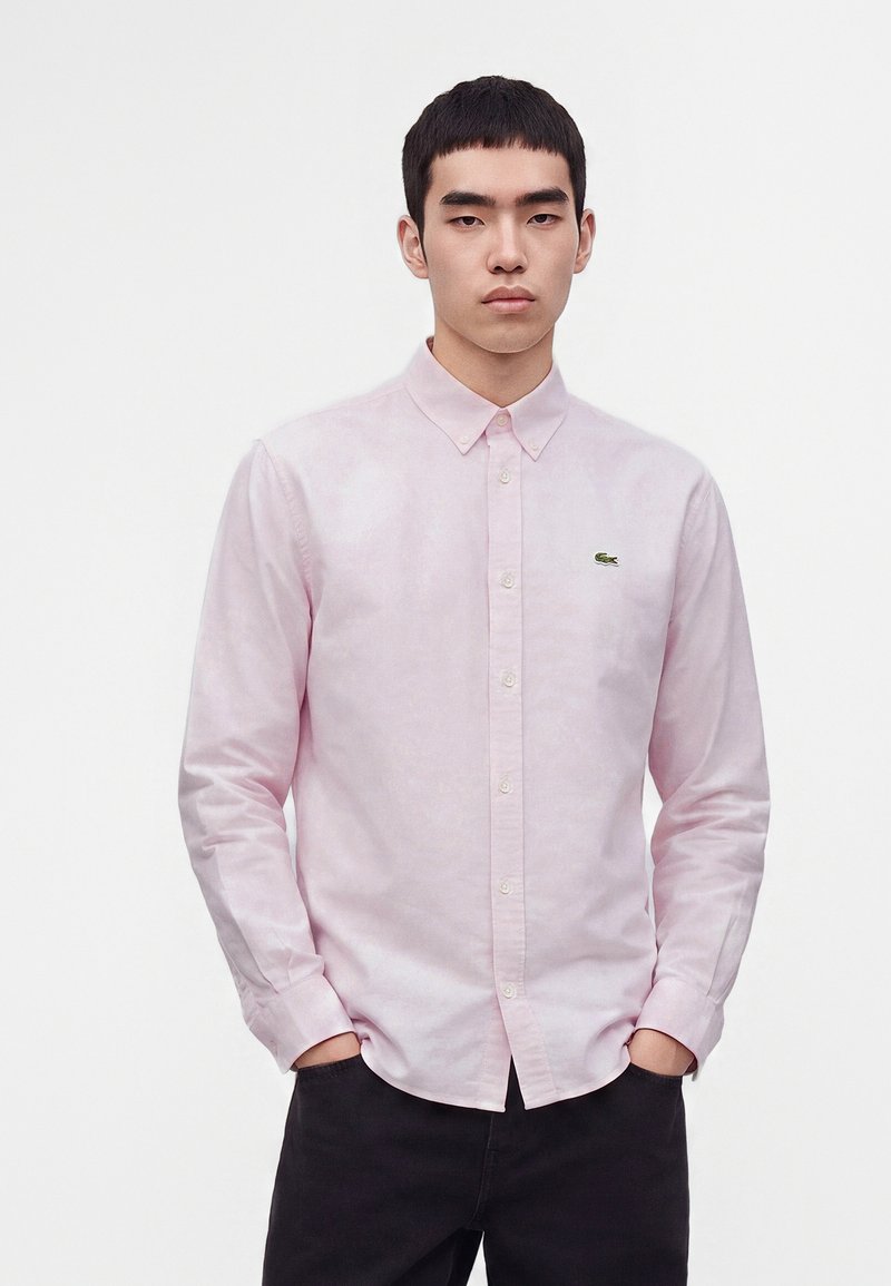 Jeune homme portant une chemise rose clair à boutons et manches longues avec un pantalon noir, debout les mains dans les poches devant un fond uni.