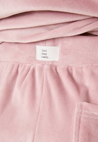 Tissu rose clair doux avec une texture lisse et moelleuse. L'étiquette indique "Cool Cosy Comfy". Présente une ceinture élastique et une poche latérale.