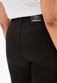 Pantalones vaqueros negros de mezclilla con cintura de altura media, corte recto, dos bolsillos traseros y una etiqueta que muestra "ARMEDANGELS" en la cinturilla.