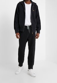 Sudadera negra con cremallera, puños acanalados y un pequeño logotipo, combinada con pantalones de chándal negros con un cordón ajustable con un diseño. Zapatillas blancas.