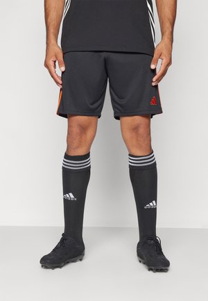 Man draagt zwarte Adidas voetbalshorts, zwarte kniehoeghoge Adidas sokken met witte strepen en zwarte voetbalschoenen, staand tegen een effen achtergrond.
